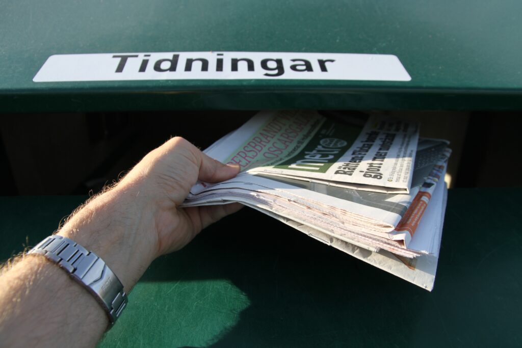 En hand sträcker in tidningar i en återvinningscontainer.