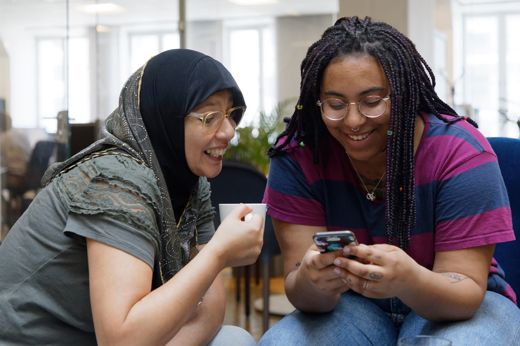 Två kvinnor, en med slöja, tittar i en mobiltelefon och ler.