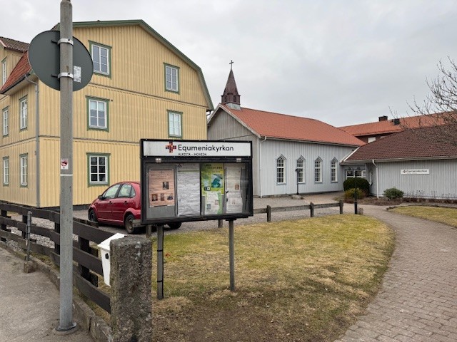 En bild av en skylt framför ett gult trähus och en vit träkyrka. På skylden står det "Equmeniakyrkan".