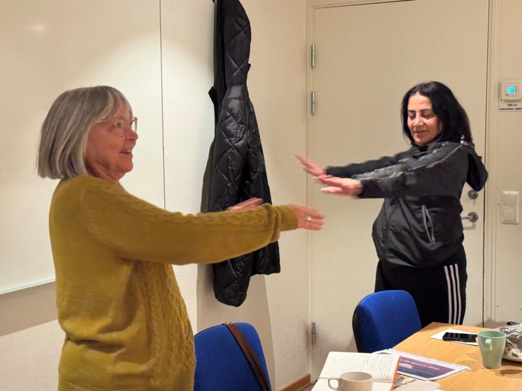 Två kvinnor i ett klassrum svänger med armarna åt sidorna. De ser glada ut.