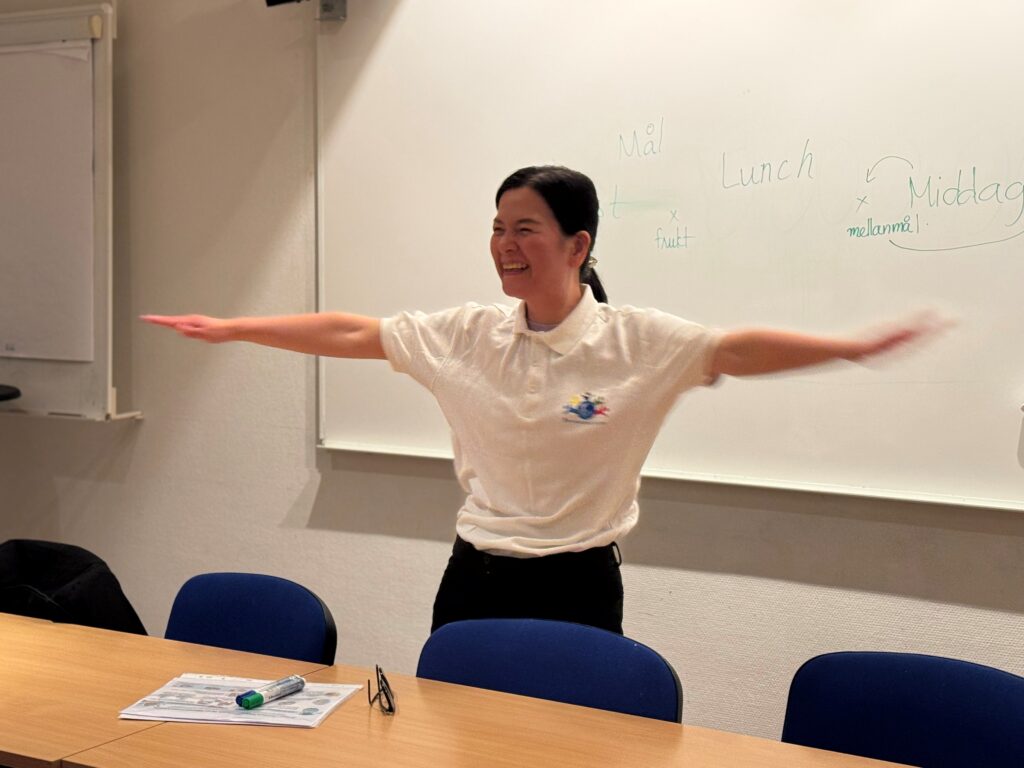 En kvinna står framför vita tavlan i ett klassrum och sträcker ut armarna åt sidan. Hon skrattar.