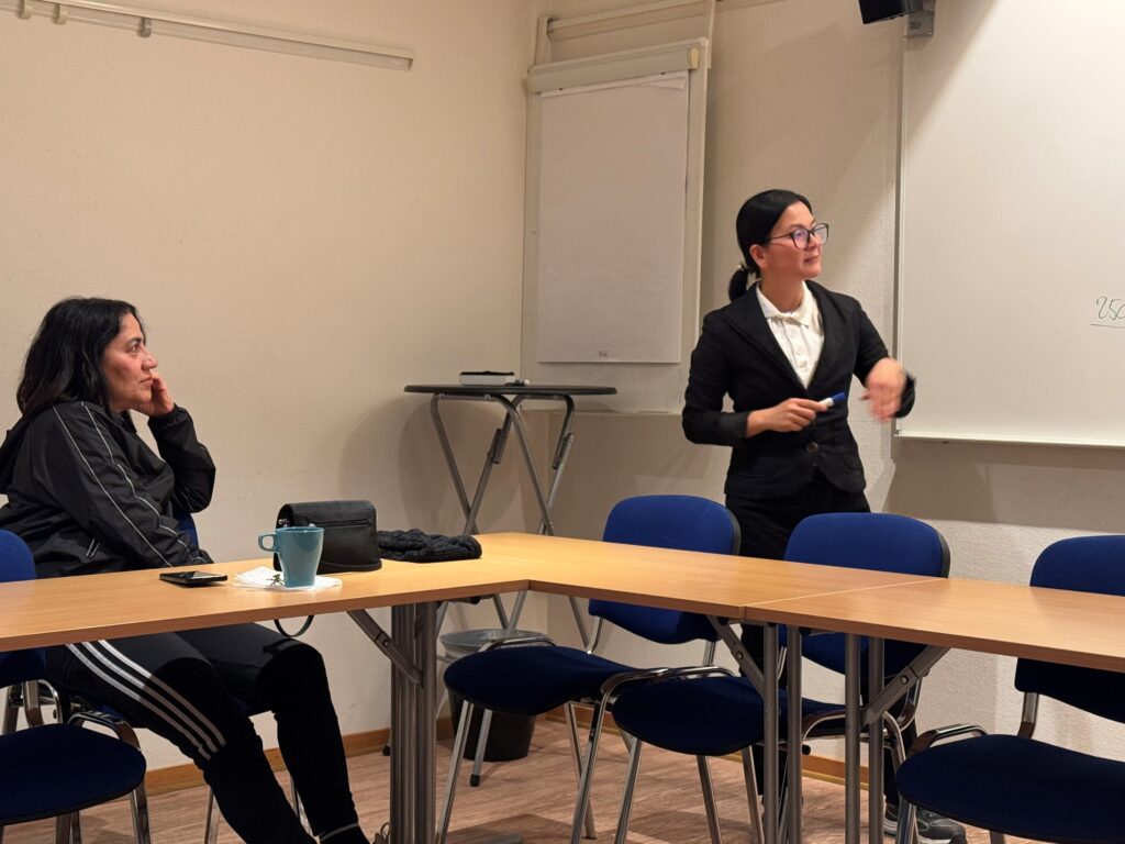 En kvinnlig deltagare sitter vid en bänk och tittar mot vita tavlan. En kvinnlig lärare står framför vita tavlan och förklarar något.