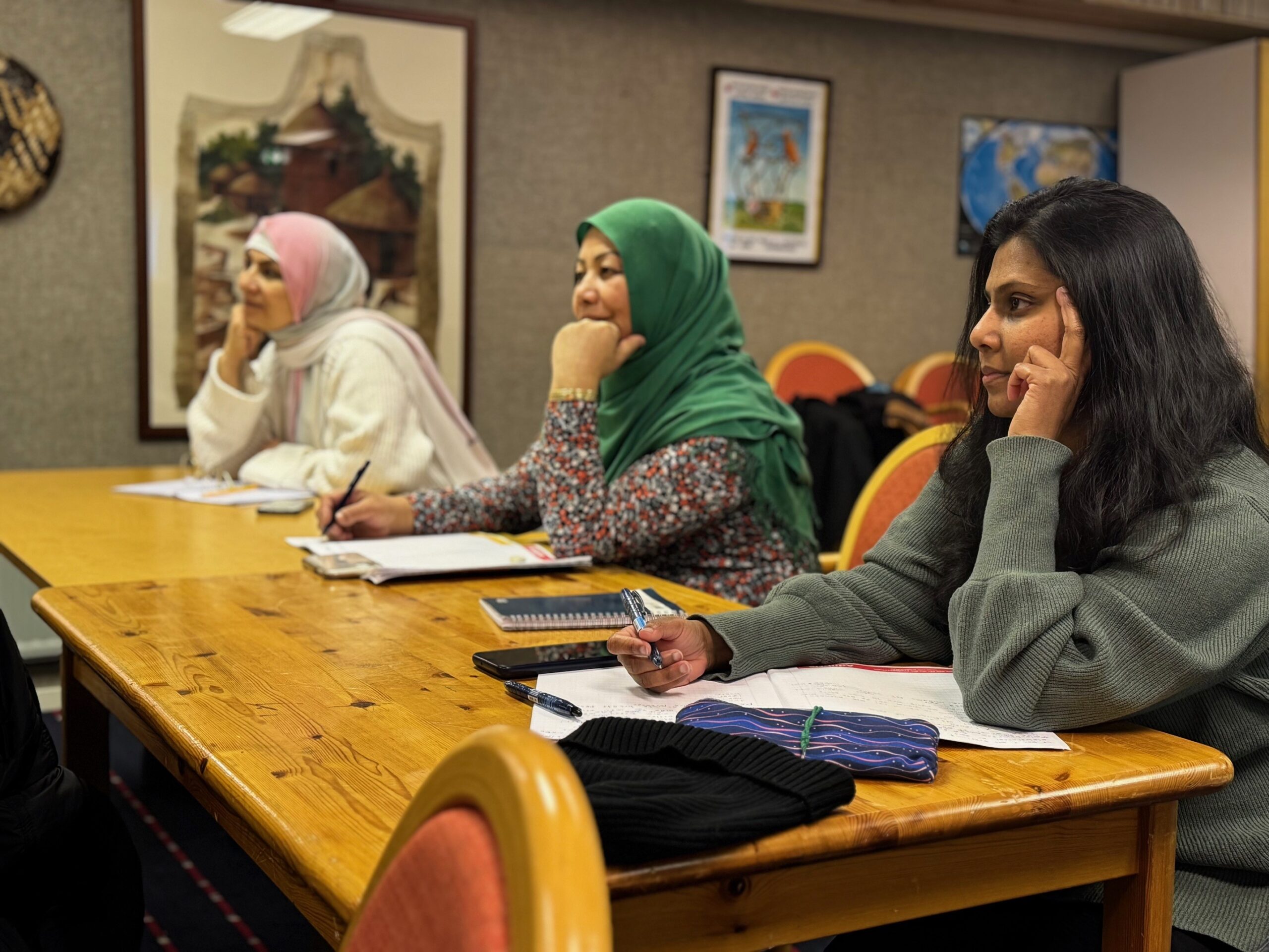 Tre kvinnliga deltagare sitter vid ett långt bord, antecknar och tittar mot läraren,