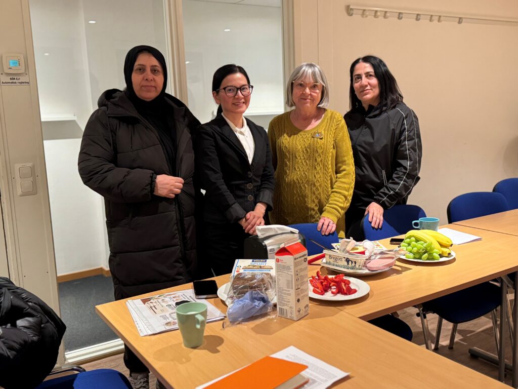 Fyra kvinnor står brevid varandra i ett klassrum. Framför dem står frukt, bröd, pålägg och mjölk framdukat.