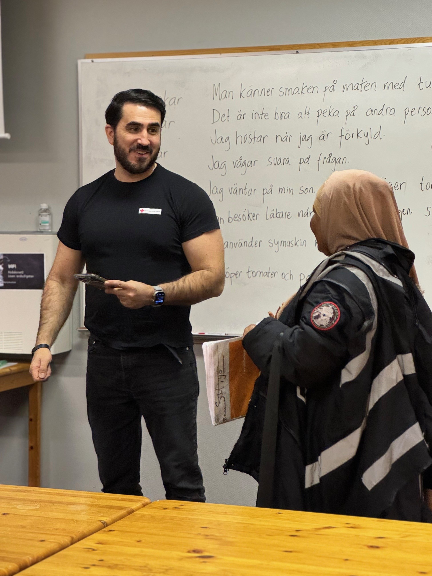 En manlig volontär pratar med en deltagande kvinna. De står upp framför vita tavlan. Han ser glad ut.