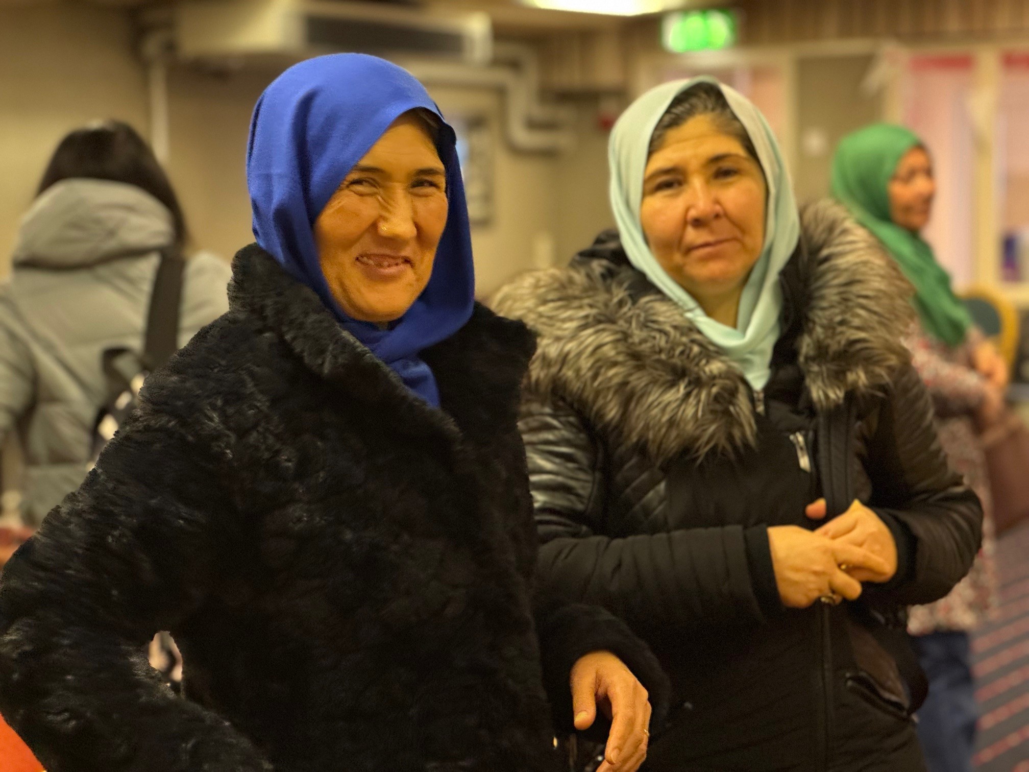 Två leende kvinnor arm i arm, med ytterkläder på, tittar in i kameran.