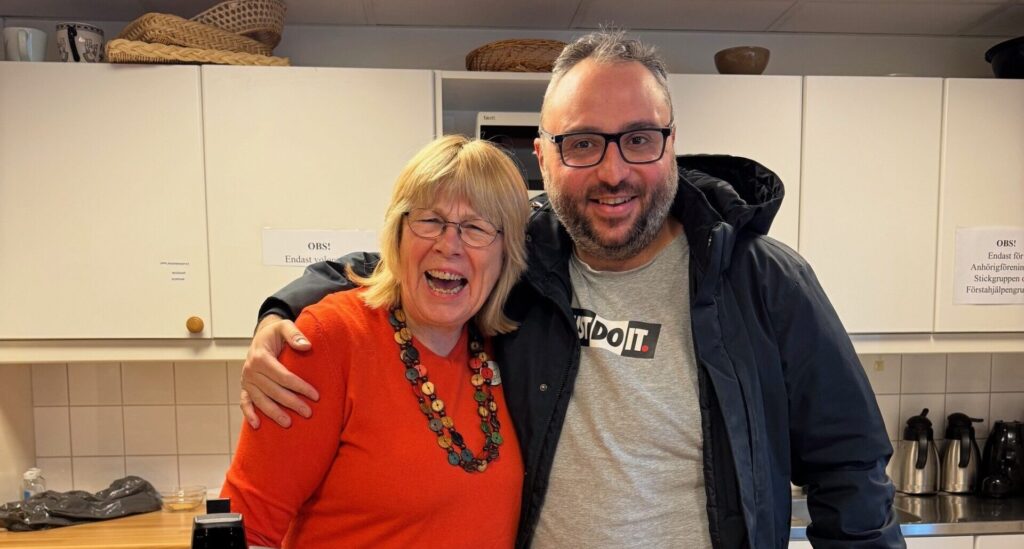 Manlig deltagare står med armen runt axeln på läraren, framför ett fikabord. De skrattar.