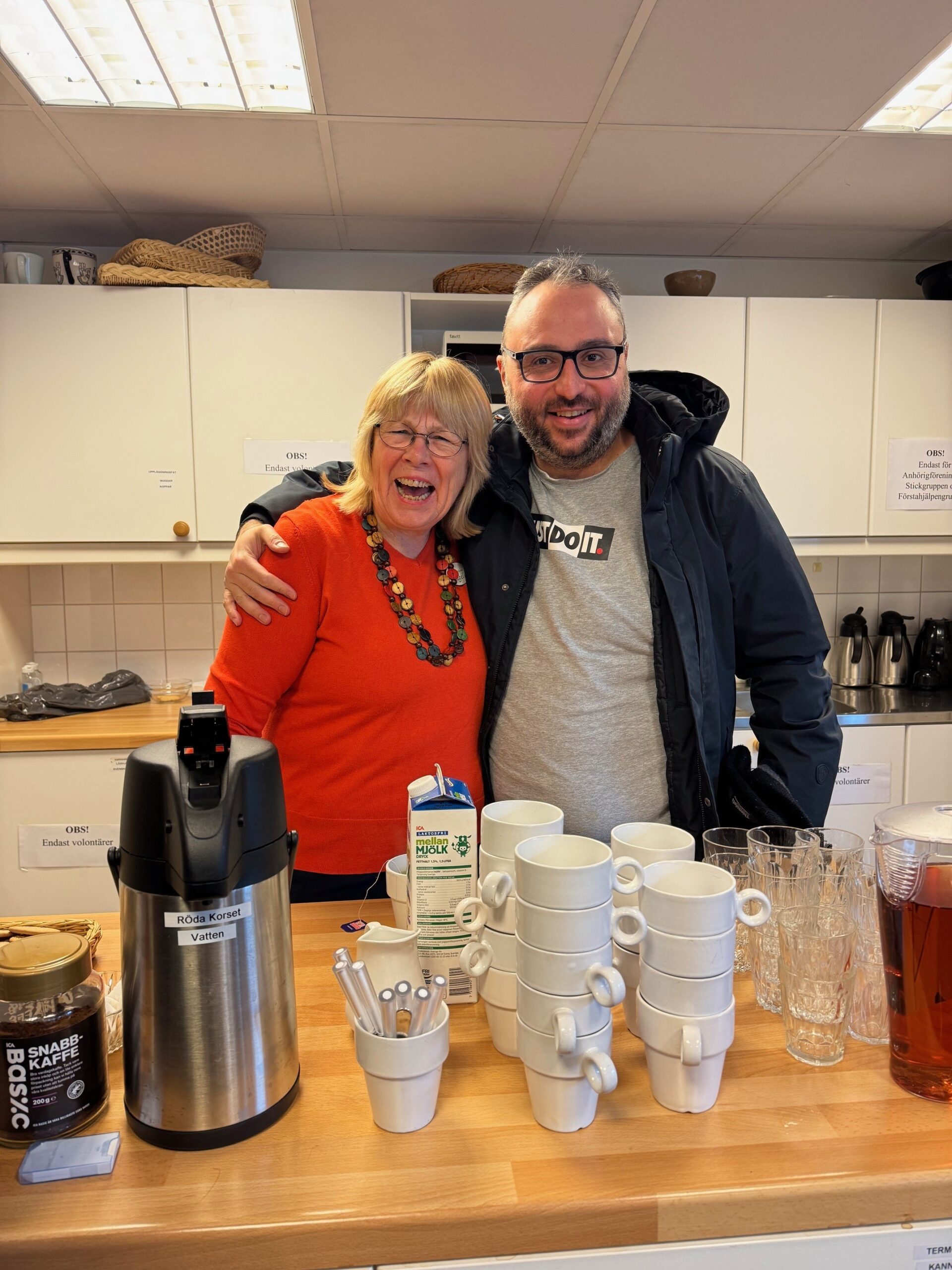 Manlig deltagare står med armen runt axeln på läraren, framför ett fikabord. De skrattar.