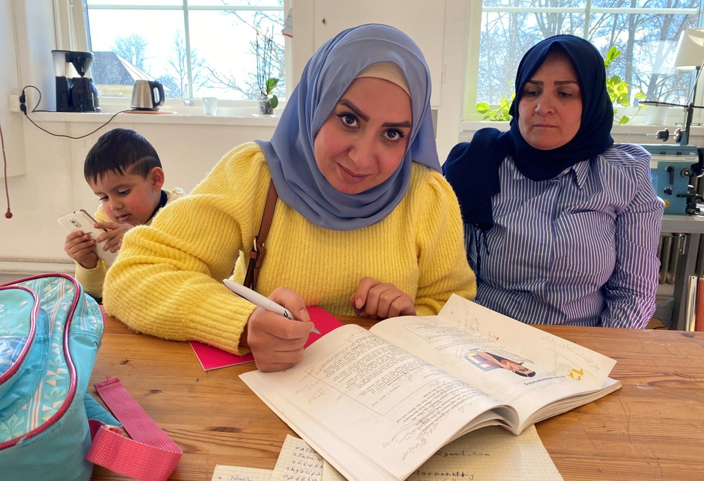 Kvinna med sjal tittar upp från en bok hon läser i. Bredvid sitter en äldre kvinna och bakom en liten pojke.