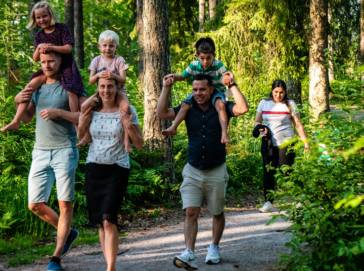 Två familjer går i skogen, barnen sitter på de vuxnas axlar.