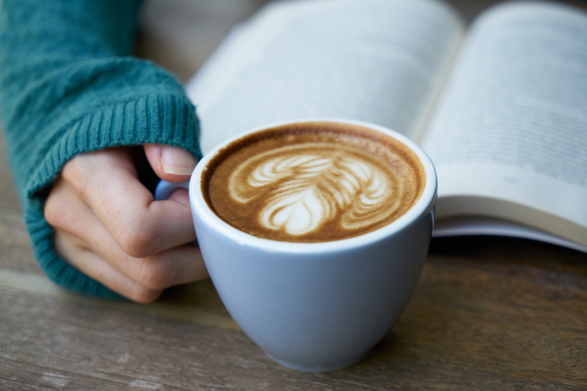 En hand håller i en kopp cappucino bredvid en uppslagen bok.