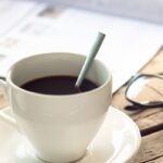 En kopp kaffe, en tidning och ett par glasögon på ett träbord.