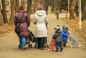 Två mammor på promenad med barnvagnar och två barn som går bredvid.