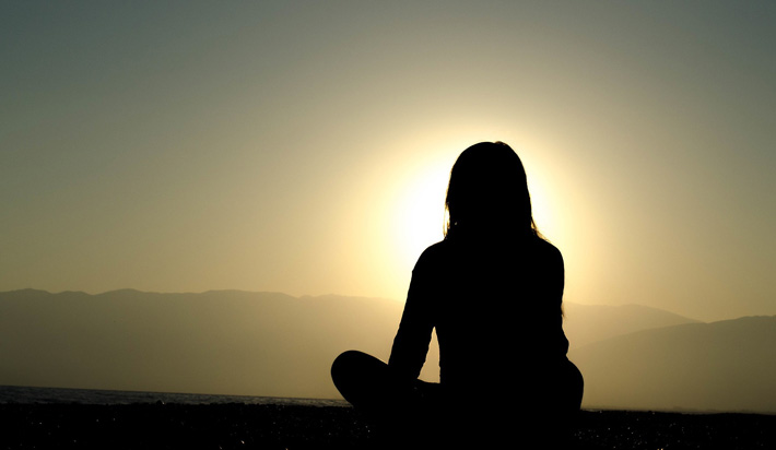 Kvinna sitter och mediterar, i siluett mot soluppgång.